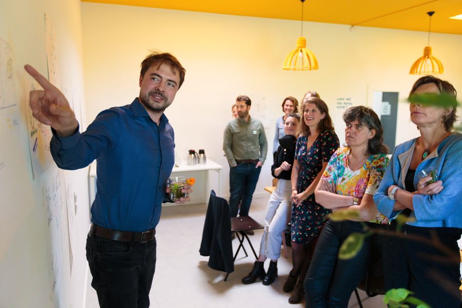 A man in a blue shirt points at drawings taped to a white wall while eight people watch and smile under yellow pendant lamps.