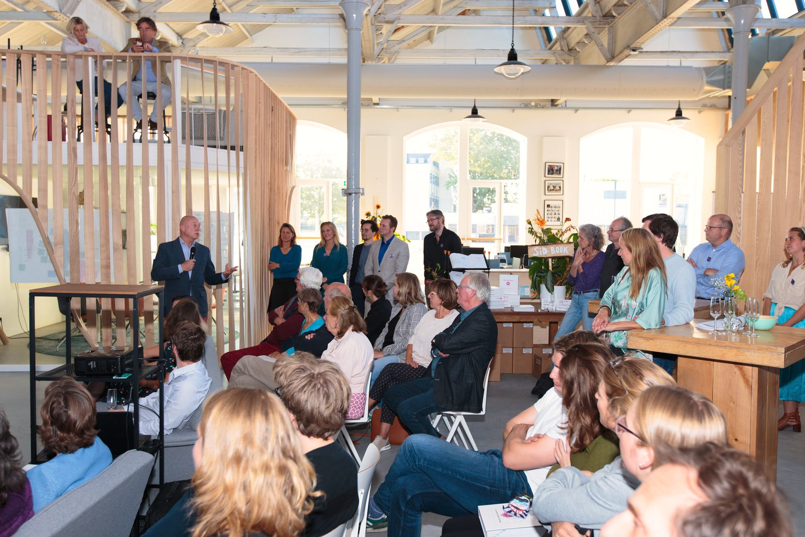 People fill the bright hall at Utrecht Community during a session, seated in rows beside a timber screen.