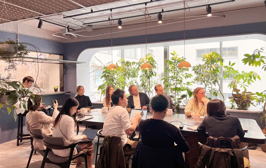 A dozen people around a long table in a plant-filled room, one presenting from a laptop while the rest listen.