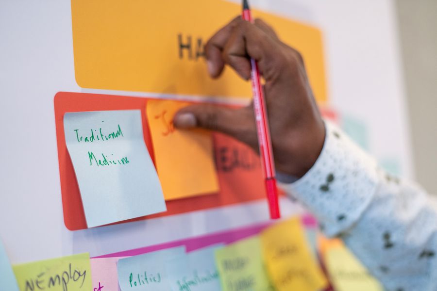 A hand holding a pink marker places a sticky note reading traditional medicine onto a board of colourful notes.