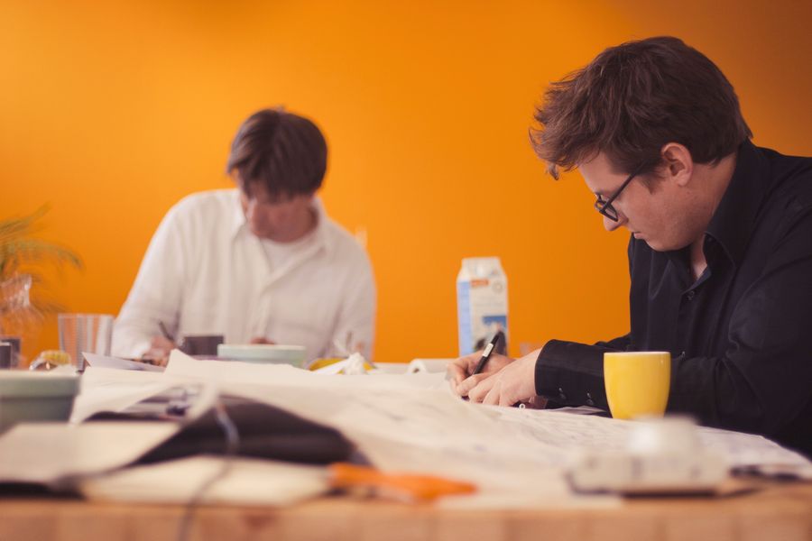 Two men work in silence over drawings spread the length of a wooden table against a warm orange wall, one writing, the other reading.