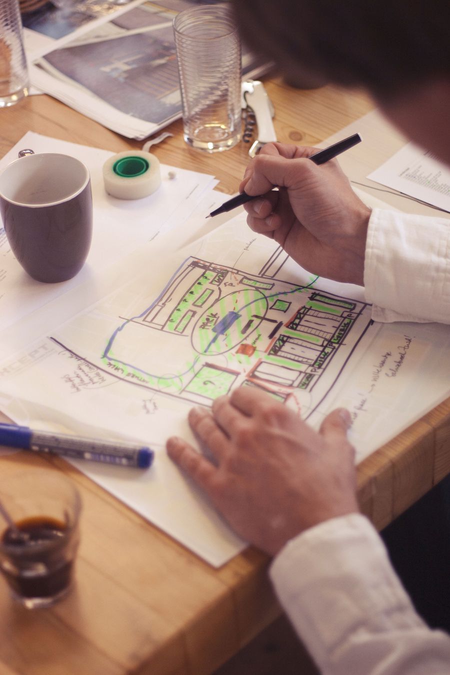 A man in a white shirt draws with a black pen on a colour marked site plan at a wooden table, a mug and a glass beside him.