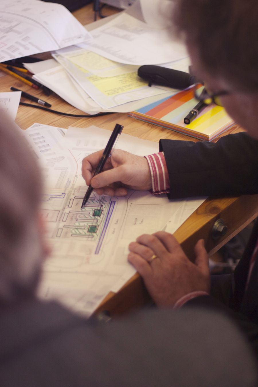 Seen over two shoulders, a man in a dark suit draws with a black pen on a printed site plan among scattered drawings and coloured paper.