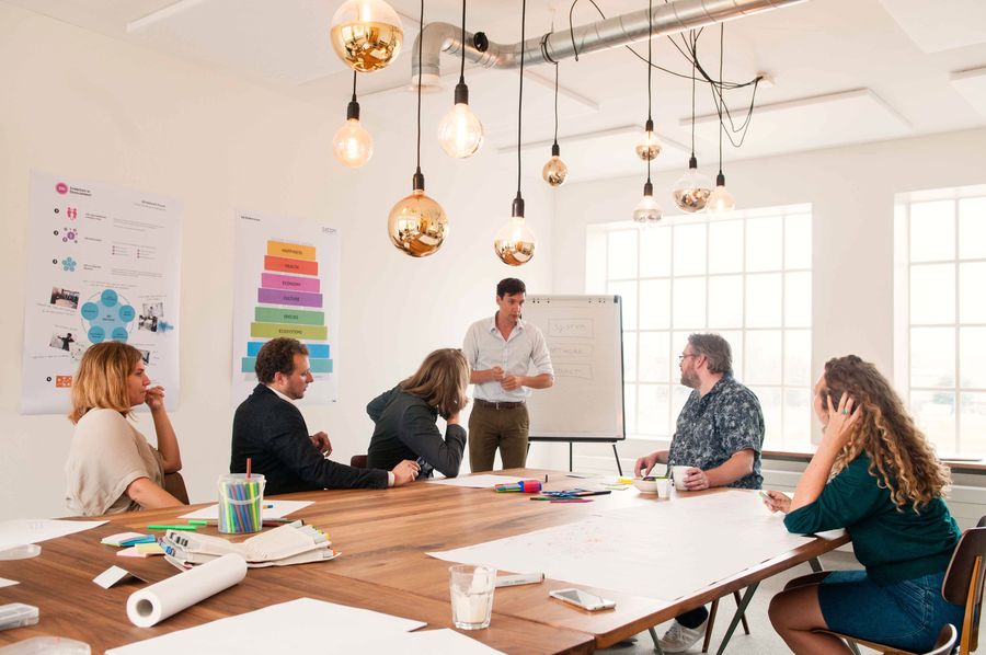 Six people work around a large wooden table in a bright white studio while a man stands at the flip chart, copper pendant bulbs overhead and markers and rolls of paper across the table.