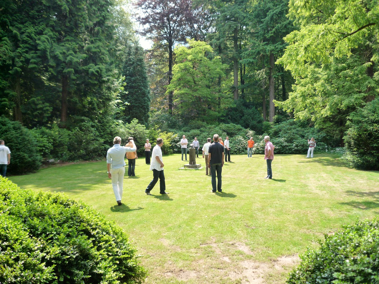 About fifteen people stand spread across a sunlit lawn ringed by tall conifers and rhododendrons, facing a small stone marker in the middle.