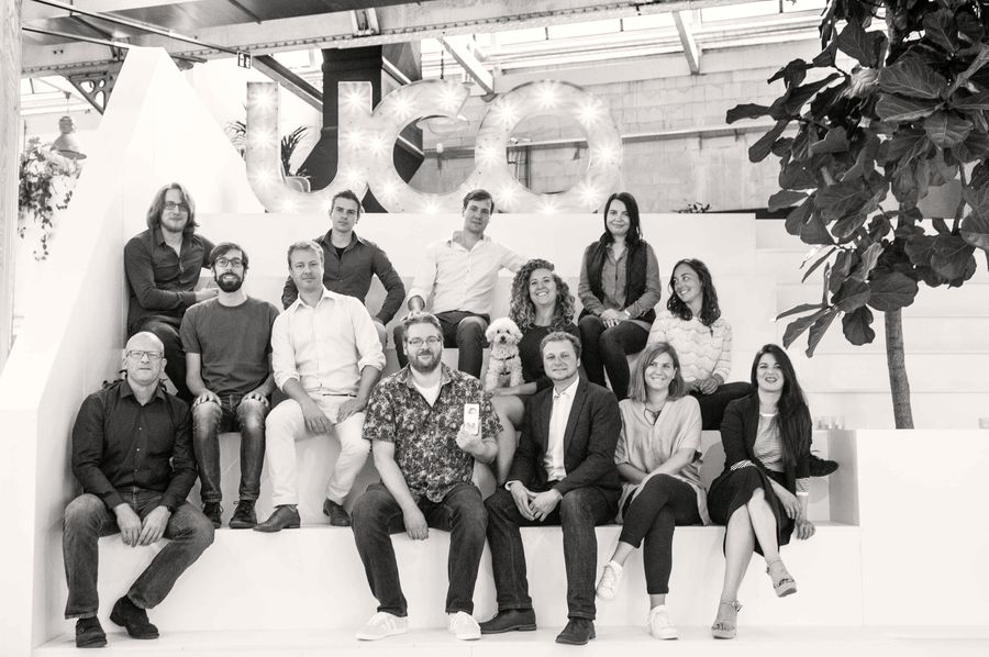 A black and white photograph of about fourteen colleagues sitting together on white steps beneath large letters spelling UCo.