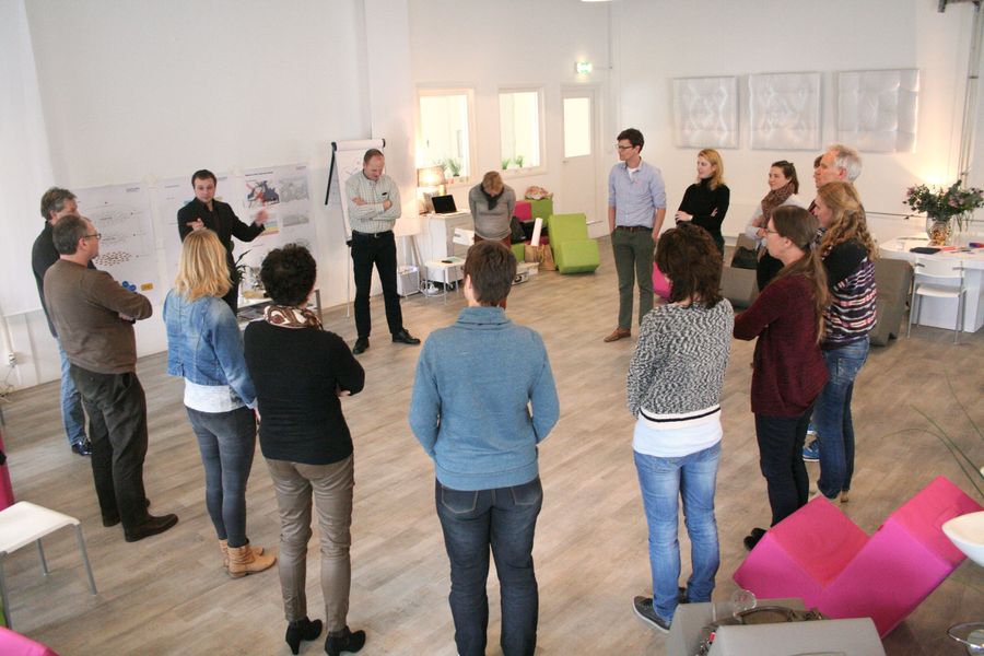 A dozen people stand in a loose circle in a bright white room while a facilitator speaks beside hand drawn posters and a flip chart.