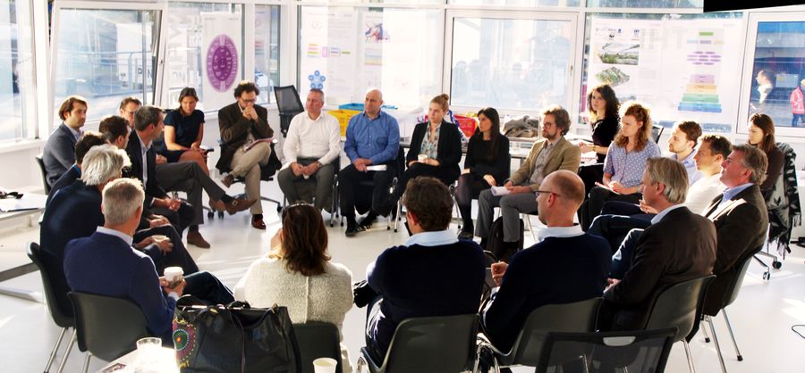 About twenty people sit in a wide circle in a bright room, project posters taped along the windows behind them.