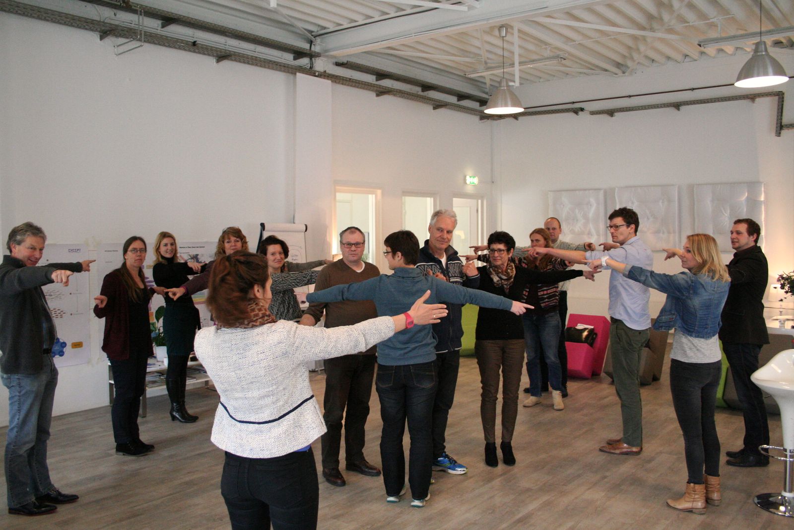 About fifteen people stand in a large circle in a white industrial room, each pointing an arm at someone else during a group exercise.
