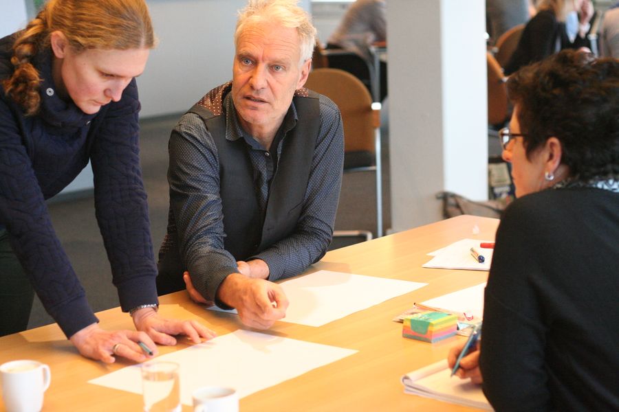 Two women and a grey haired man talk over blank paper at a light wood table, one woman leaning in on her hands.