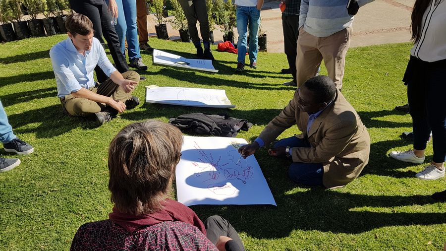 A group sits on a sunlit lawn around large hand drawn system maps, one man kneeling to write while another sits cross legged watching.