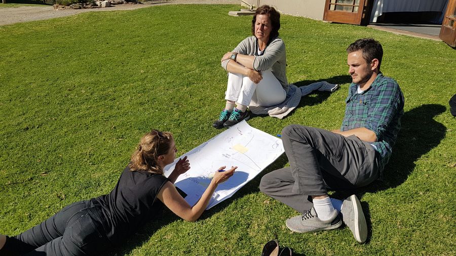 Three people sit and lie on the grass around a hand drawn map in bright sun, one propped on her elbows explaining to the other two.