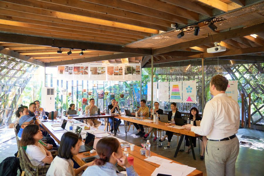 About fifteen people sit along two long wooden tables under a bamboo and timber roof, watching a man present beside framework posters, tropical planting visible through the glass walls.