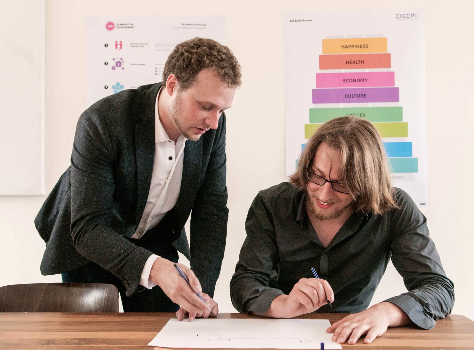 Two colleagues lean over a large sheet of paper at a table, the framework posters pinned to the wall behind them.