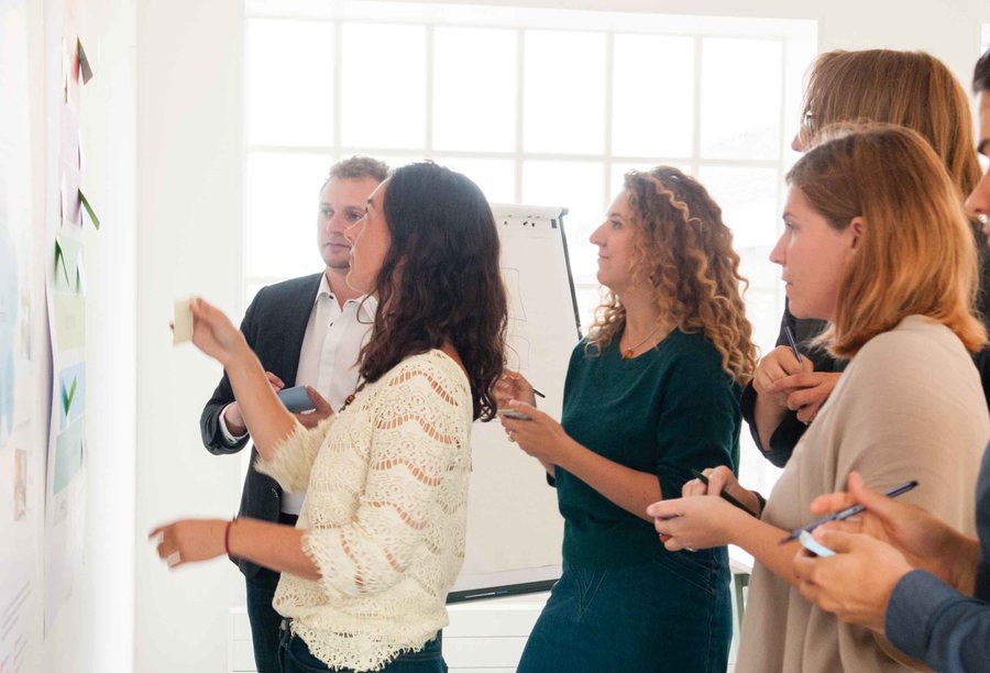 Five people stand at a studio wall, one placing a card on it while the others watch and talk.