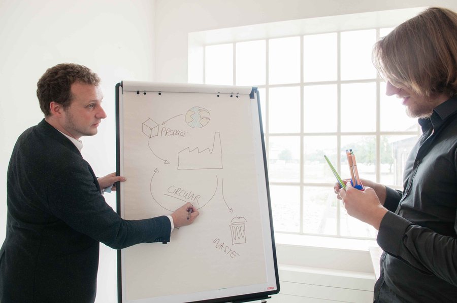 A man draws a diagram on a flip chart while a colleague watches, pen in hand.