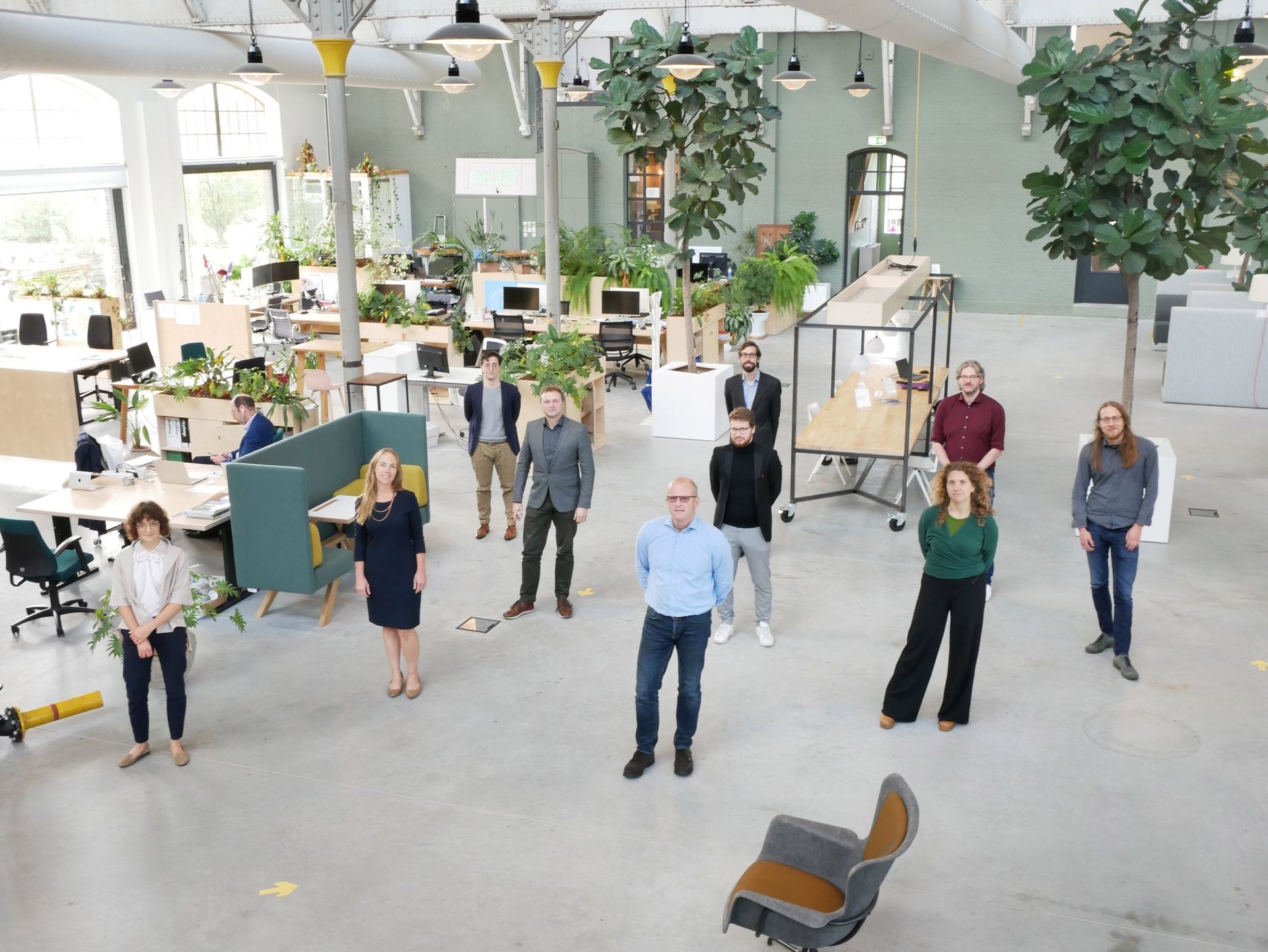 A wide studio seen from above, about fifteen people standing in ones and twos among plants, desks and workbenches.