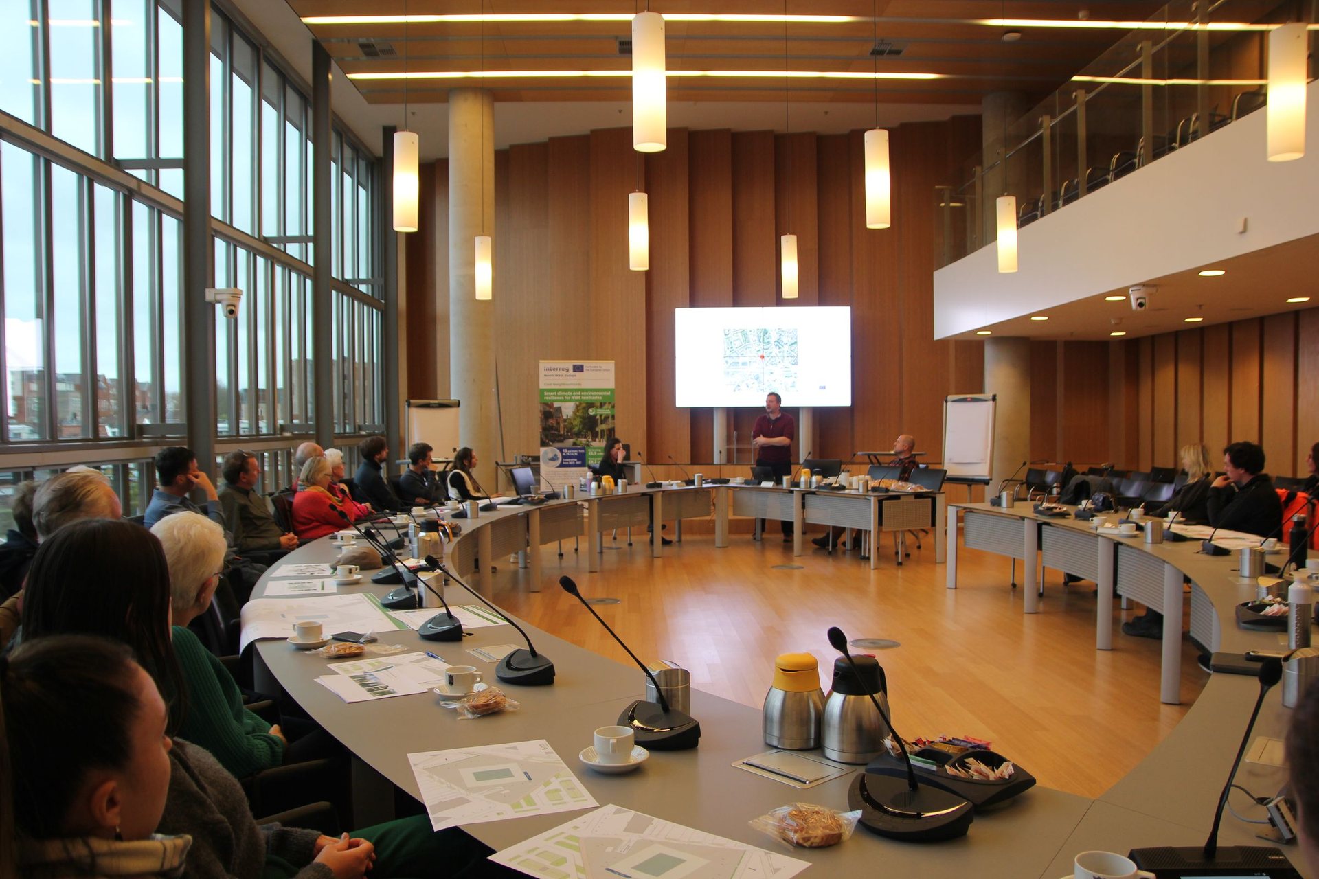 A wide arc of tables with about thirty people seated at microphones, a presentation on screen and daylight through the glazed wall behind.
