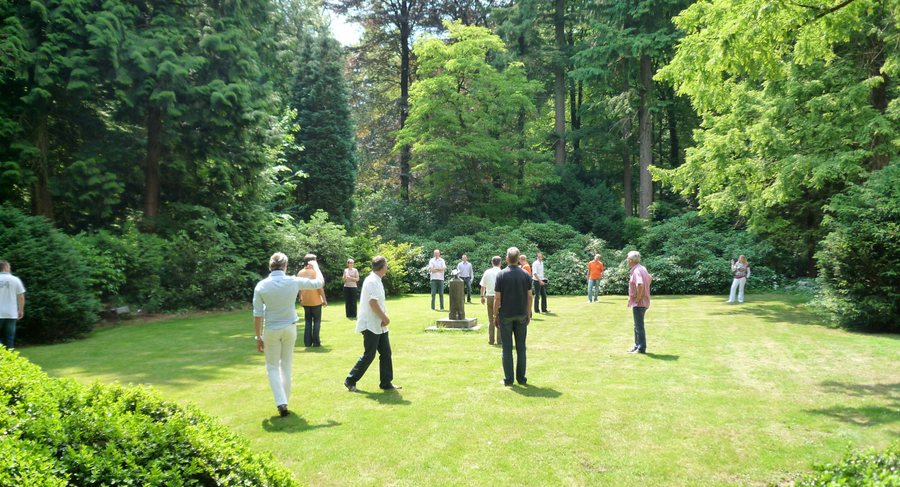 A group of about fifteen people stands together in a sunlit clearing ringed by tall trees.