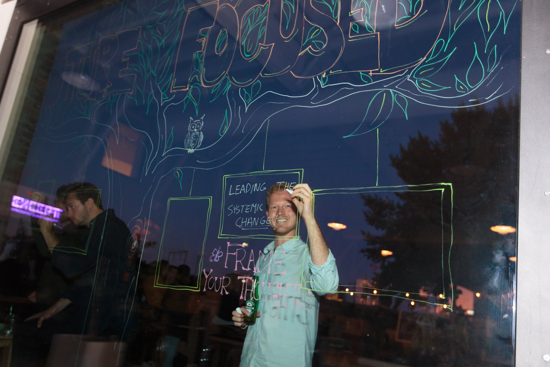 A man stands behind a large window drawn on in coloured marker, the words leading the systemic change and reframe your thoughts among painted leaves.