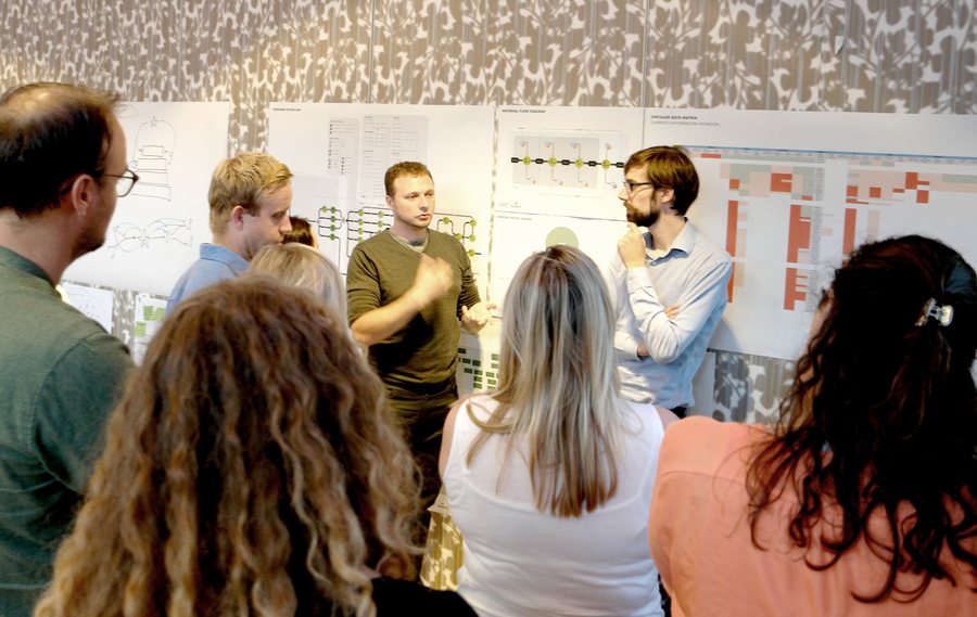 A group stands in front of a wall of printed diagrams and timelines, talking in twos and threes.
