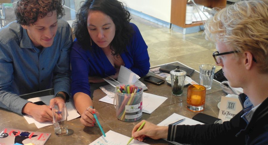 Three people work on paper cards at a table, a pot of coloured pens between them.